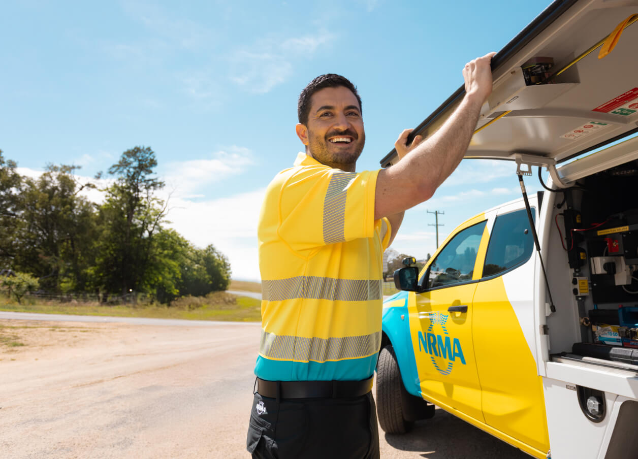 A man dressed in an NRMA uniform stands by a roadside service vehicle, smiling while the side trunk is open.
