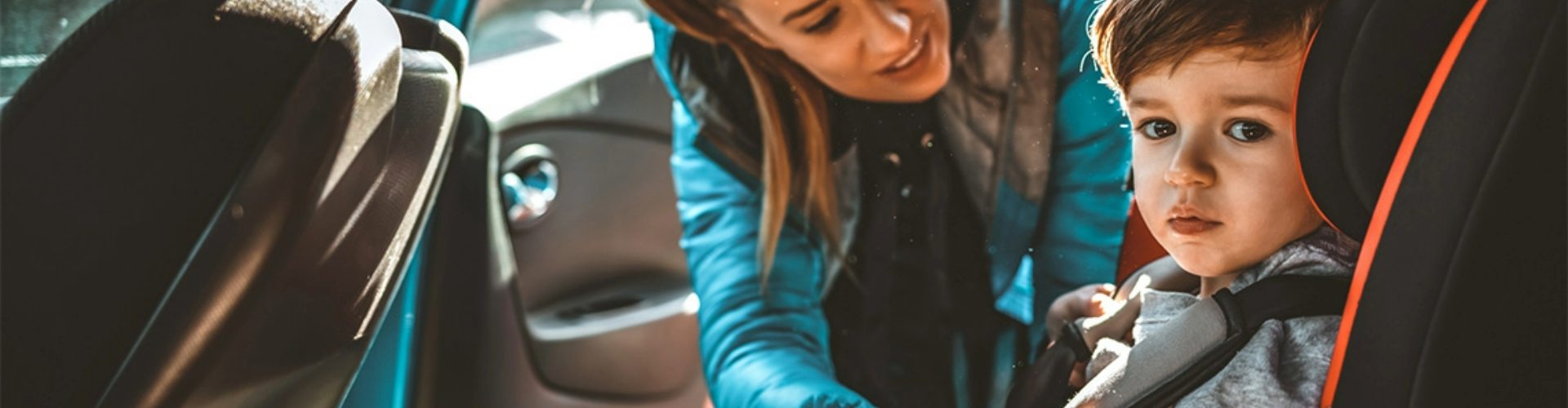 mother-child-car-seat-restraint-1920x500 A mother leaning into a car to secure her toddler's child seat restraints.