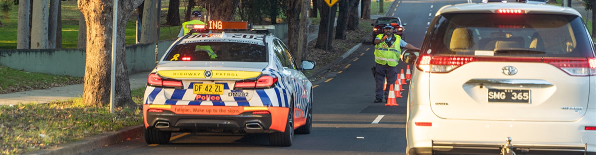 police-running-drug-stop-1920x500 A policeman puts his hand out to slow cars passing by a row of pylons set up to test drivers for substance use.