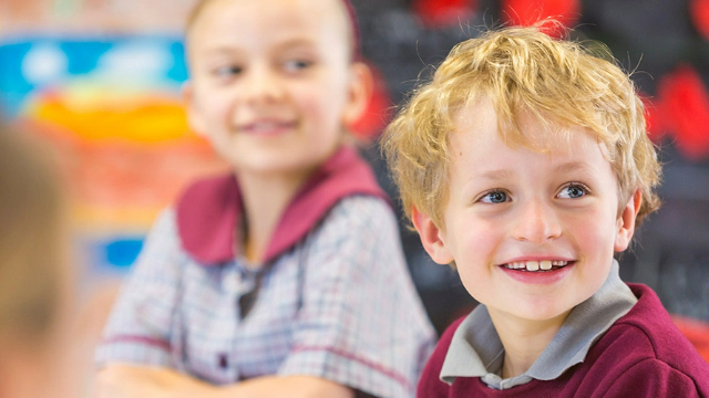 A boy and girl sitting in a classroom, turned towards the camera and smiling. 