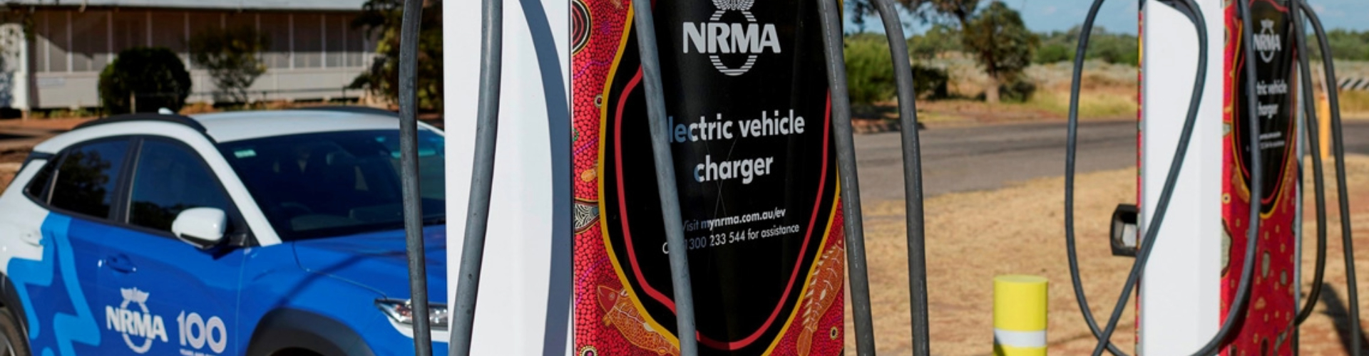 Two EV charging stations, decorated with Aboriginal art dot designs, along a sunny, rural road. 
