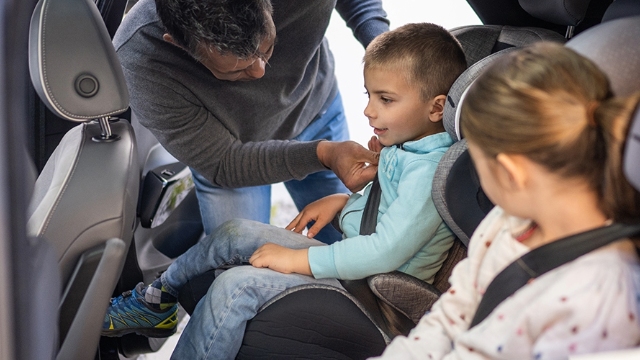 male child being secured in their car seat by their dad while their sister watches