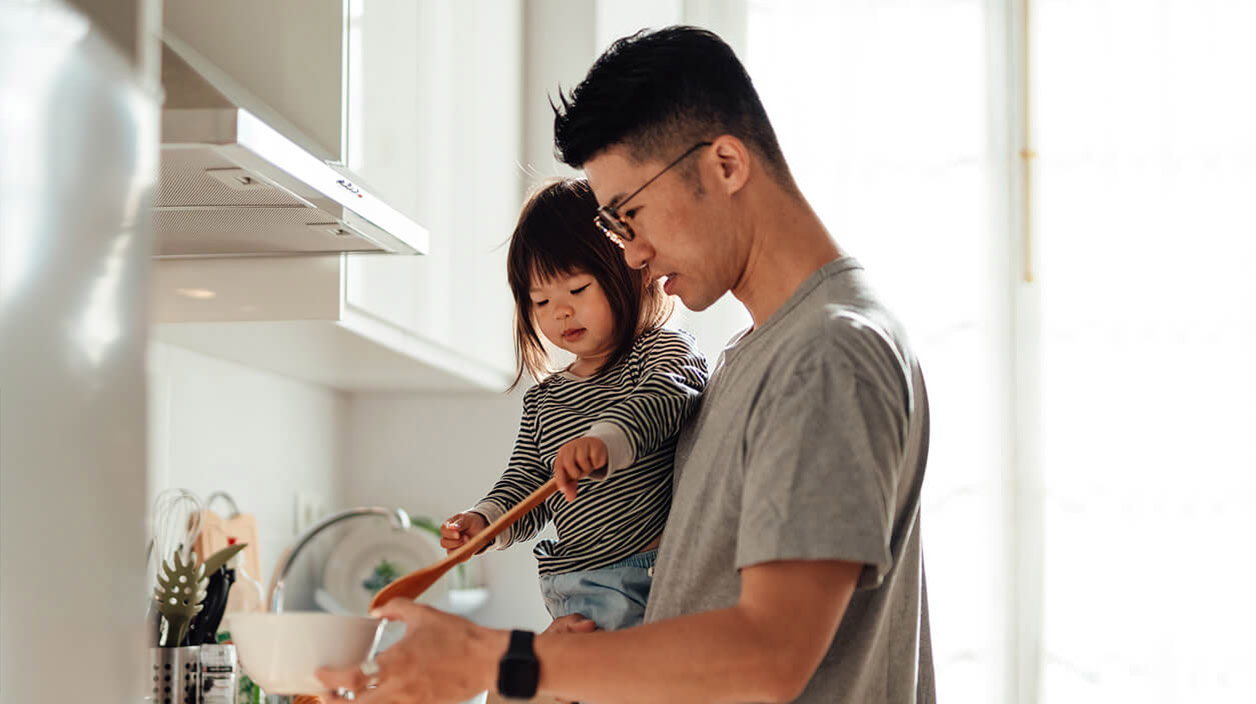 A father holding their child while cooking together at a kitchen stove, with the child stirring food in a bowl.