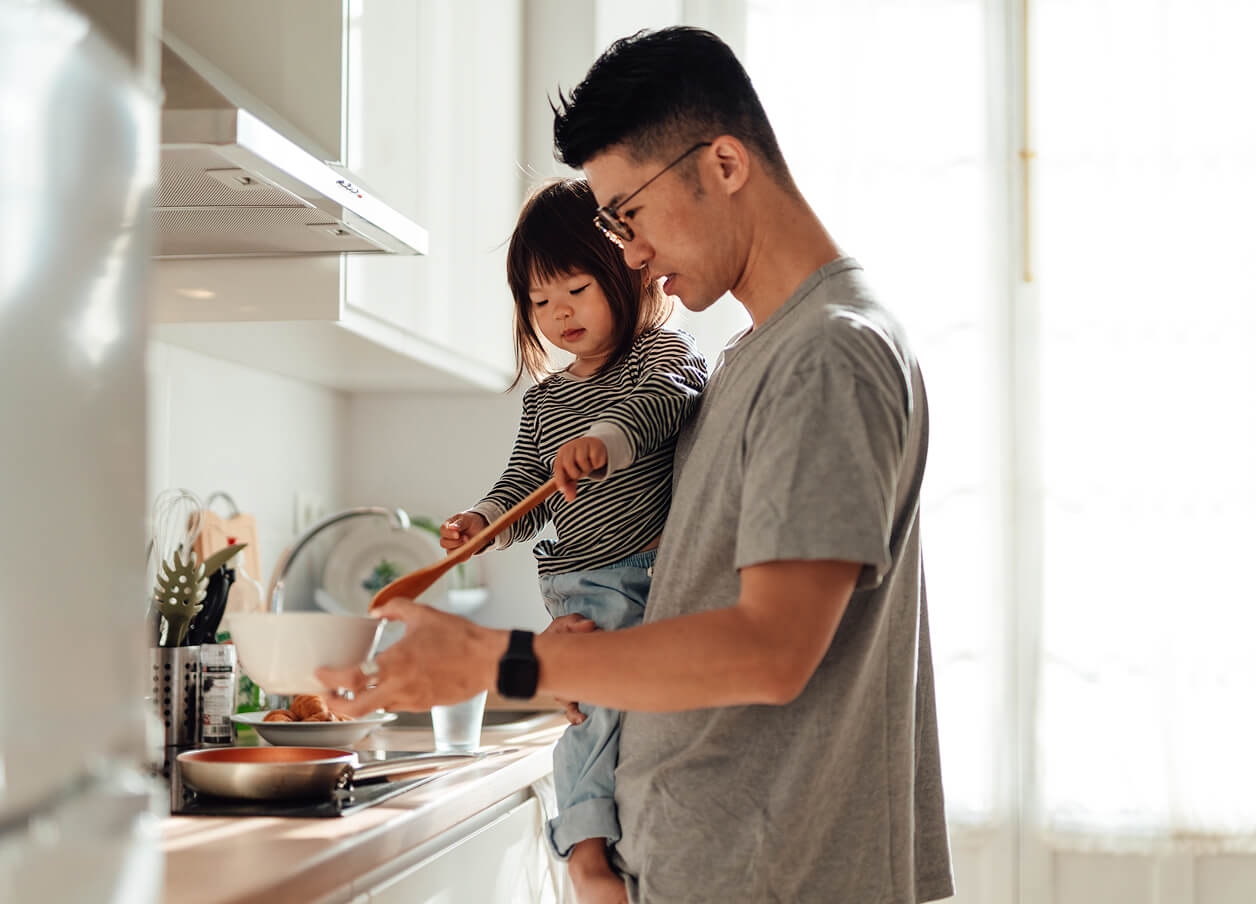 A father holding their child while cooking together at a kitchen stove, with the child stirring food in a bowl.
