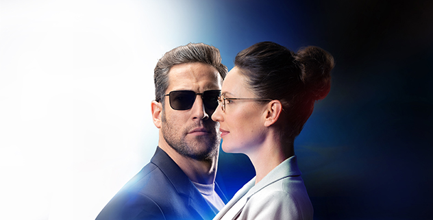 A man wearing dark sunglasses faces forward while a woman in clear glasses stands in profile beside him; the background is split into light and dark halves, suggesting contrasting perspectives or styles.