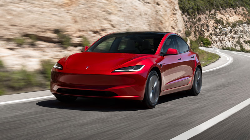 A red Tesla Model 3 driving on a road next to a rock wall