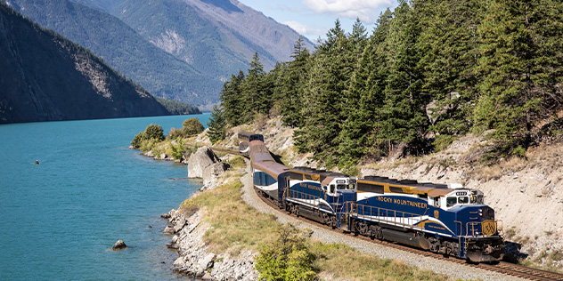 A train on the rails next to a crystal lake surrounded by mountains