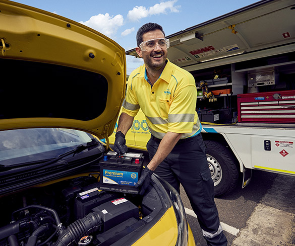 A Roadside assistance NRMA staff member in uniform holding a car battery in his hands smiling while standing next to a vehicle with an open bonnet