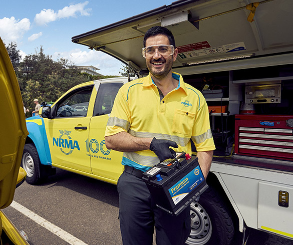 A Roadside assistance NRMA staff member in uniform smiling while holding a large battery in his hands, showcasing its size and importance.