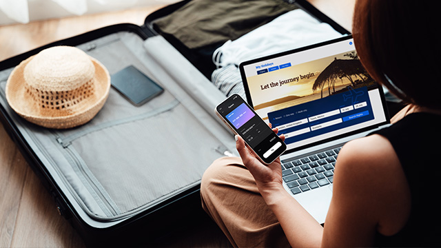 Over the shoulder view of young woman booking a flight via laptop and smartphone for vacation