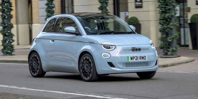 Front and side-angle view of a powder blue Fiat 500e driving along a city road with a building in the backdrop.