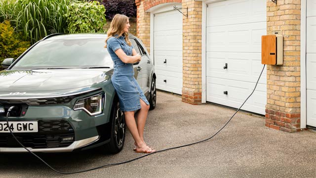 A woman in a denim dress leans on a blue Kia Niro EV while it charges next to a brick house