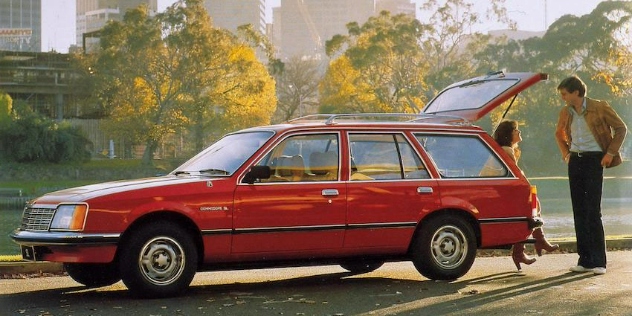 Side View of a red 1978 VB Commodore, with a lady sitting at the edge of the boot and a man standing in front of her.