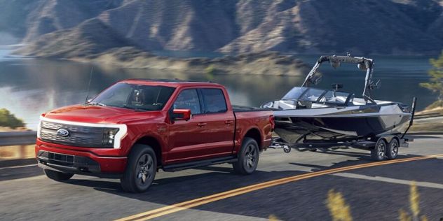 A red Ford F150 Lightening electric ute tows a boat up a lakeside road