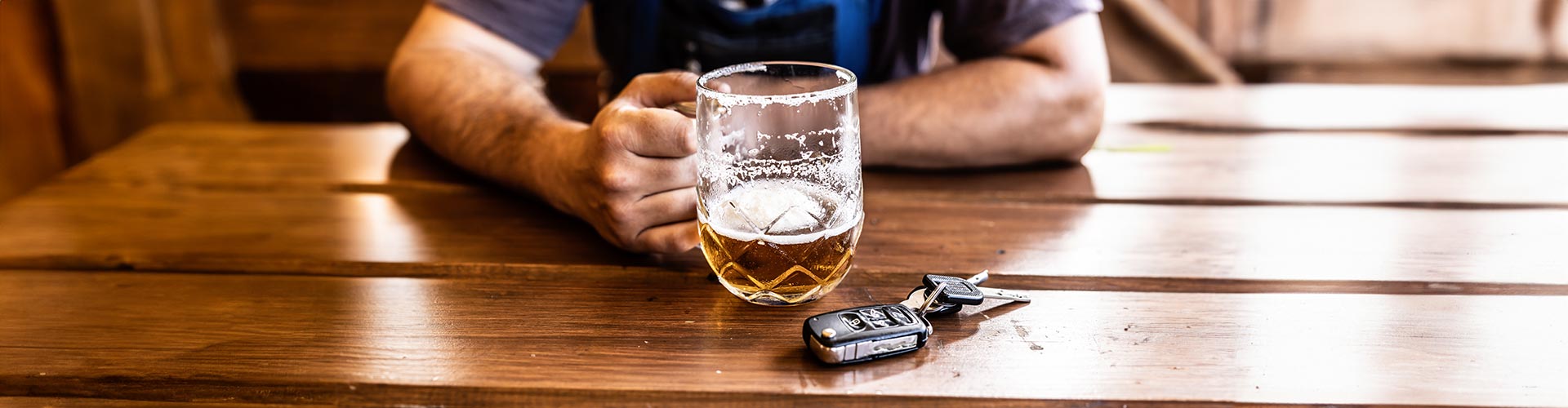 A hand of a man holding a beer on a wooden table next to s set of car keys