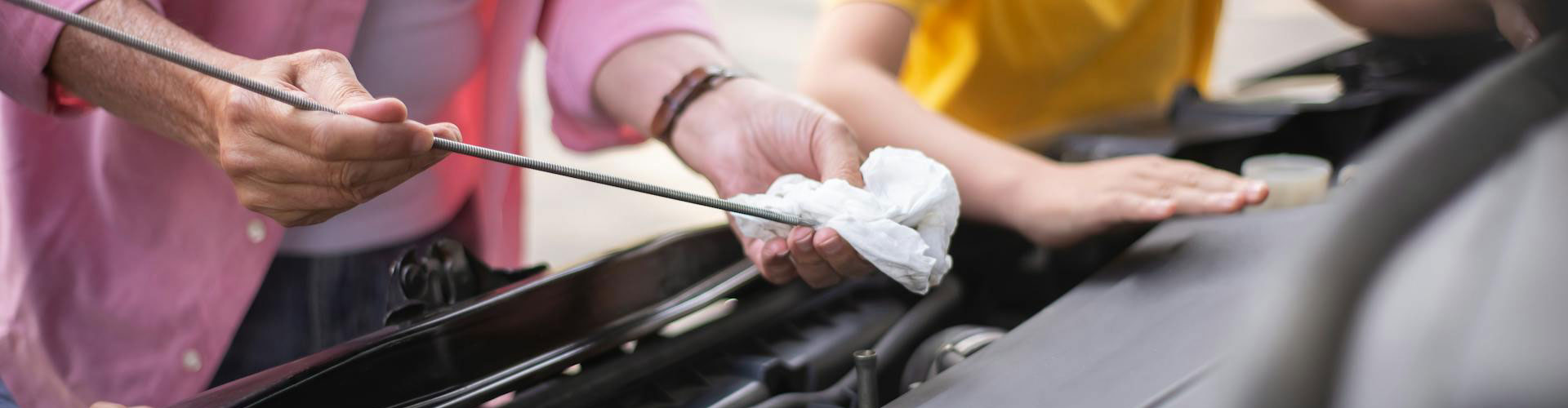 A hand of a father in a pink sleeve can be seen wiping the oil checker of a car next to a child in yellow shirt