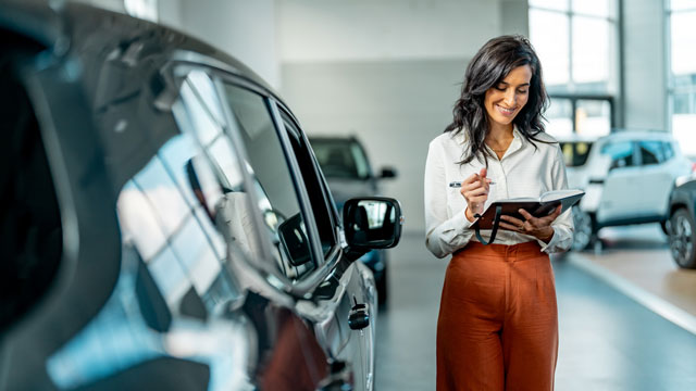 Car dealer with long hair wearing orange pants in showroom looking at a ledger book