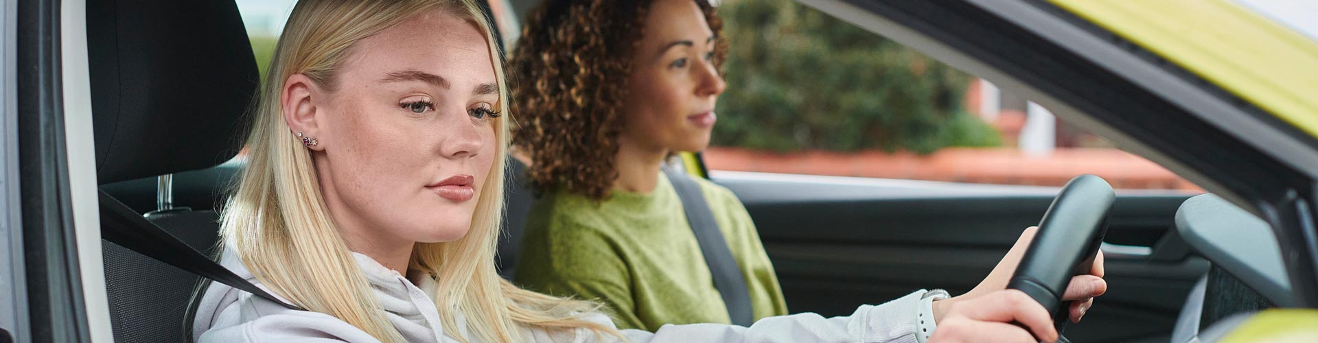 Young woman with long blonde hair in yellow car checks her rear vision mirror while her mother looks head