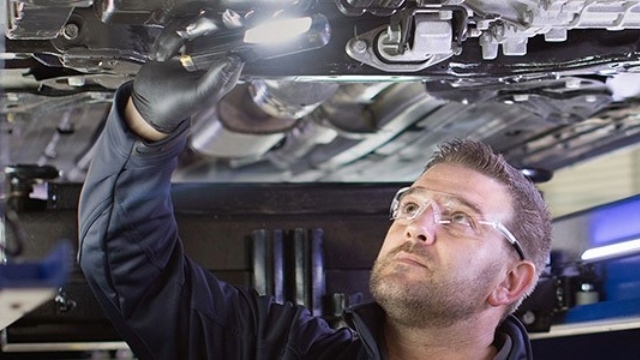Motorserve Mechanic motor mechanic stands under a car raised on a hoist and holds a light to inspect the underside