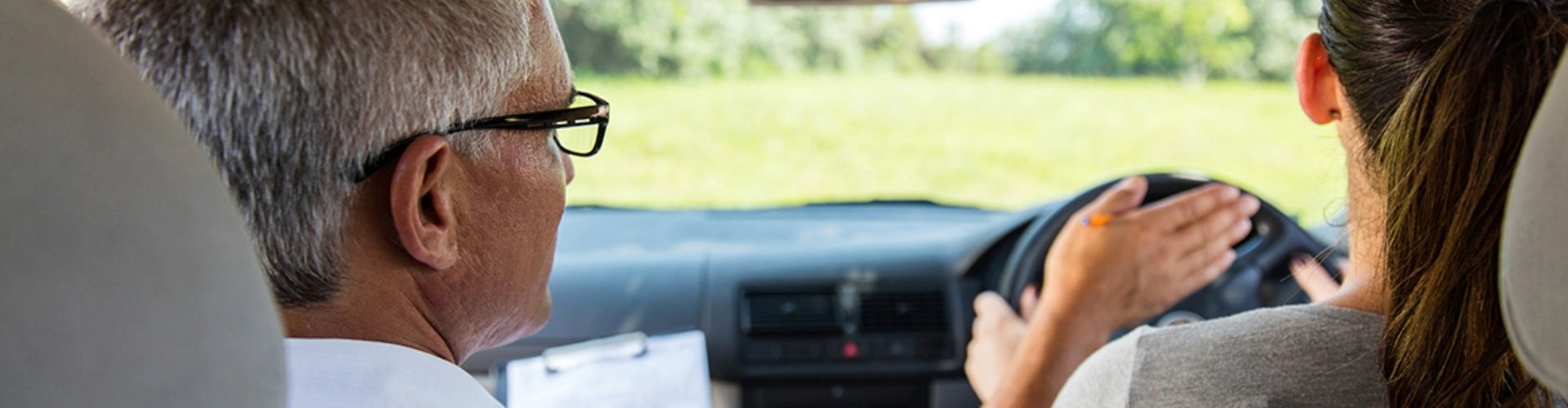 Teaching someone to drive a man with grey hair and glasses in the passenger seat gestures right to a young woman with long hair who is driving the vehicle