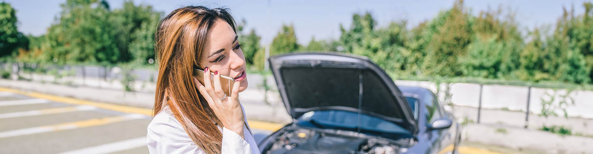 A woman calls for help on her smartphone in front of her broken down car with bonnet up