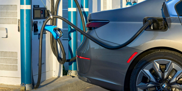 The back of a grey electric car, with it's charge port open and plugged into a charging station on a sunny day. 