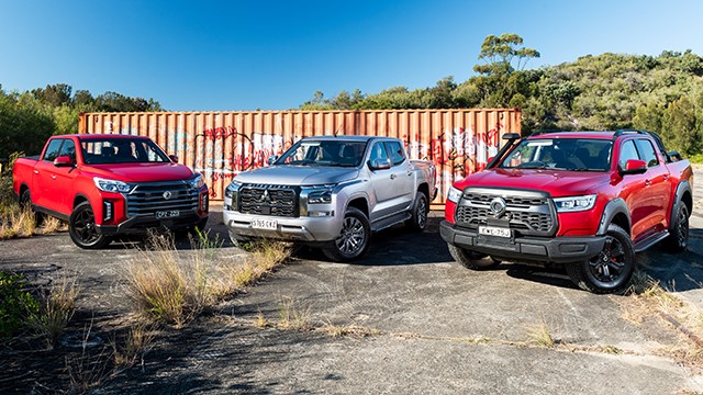 Three light trucks, two red and one silver, parked in a sunny, overgrown lot.