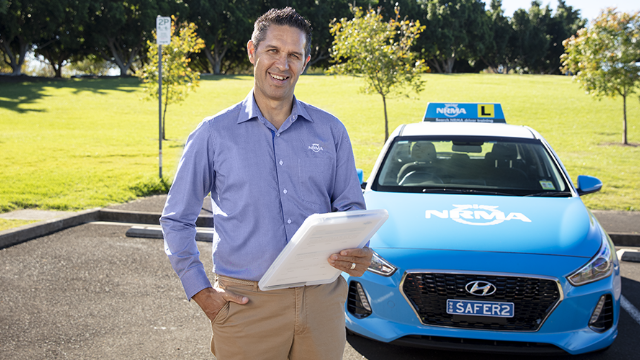 An NRMA driving instructor standing in front of a parked NRMA driving instruction vehicle