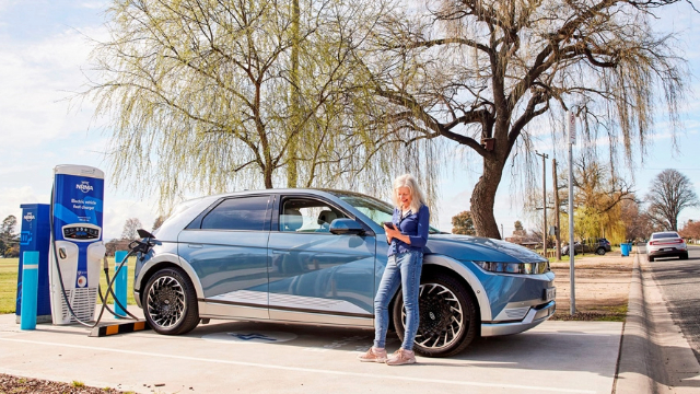 Someone leaning against their EV and looking at their phone while it charges at a public charge station.  