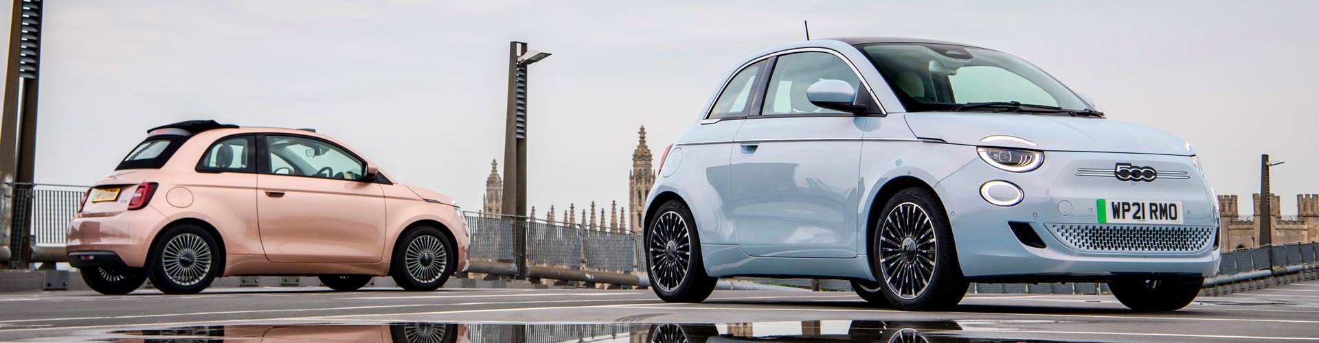 A pink and a light blue 2023 Fiat 500e parked on a bridge in London