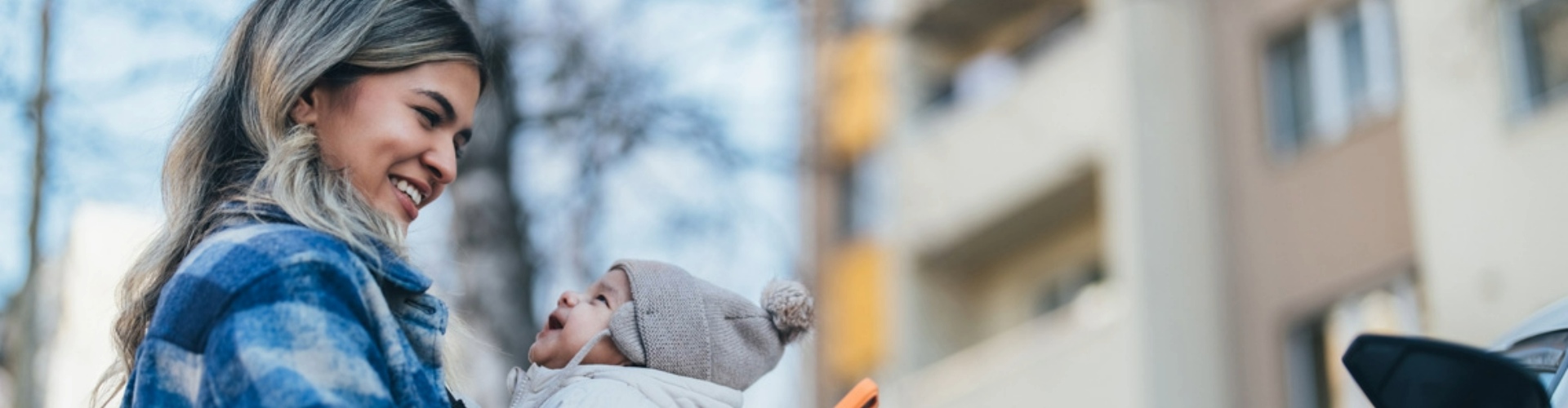 A young woman wearing a blue and white plaid shirt, smiling while holding her phone in one hand and her baby with the other.