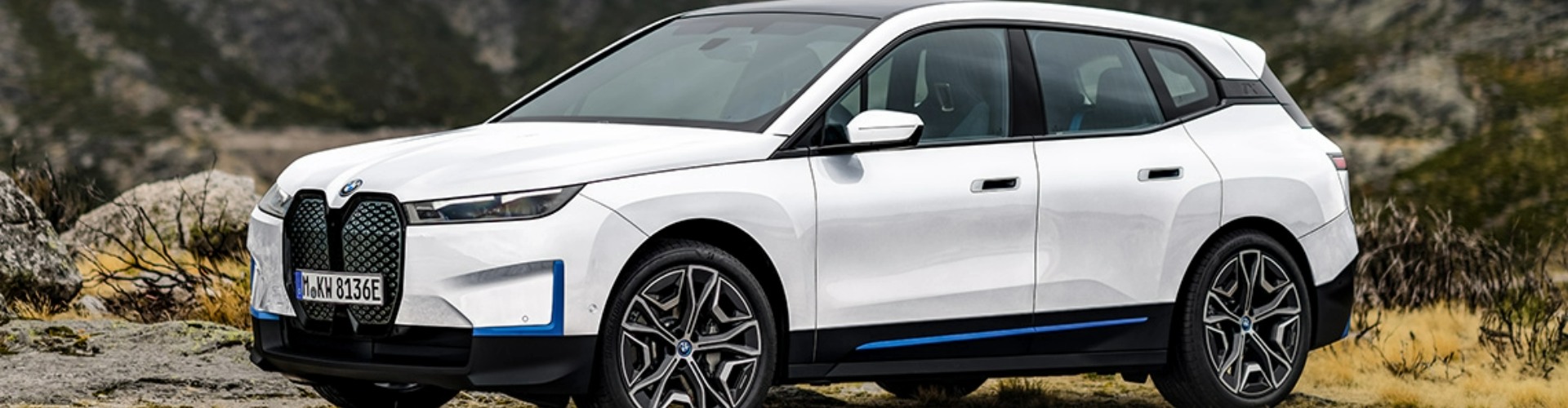 Side-angle view of a white 2022 BMW iX parked in outdoor terrain with a mountain backdrop.