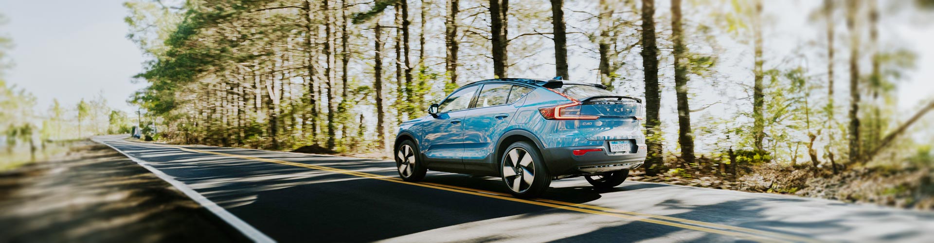 A blue Volvo C40 speeds along a road lined by pine trees