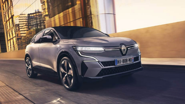 Front-angle view of a brown Renault Megane E-Tech travelling along a bridge with a large building that has mirrored windows in the background.