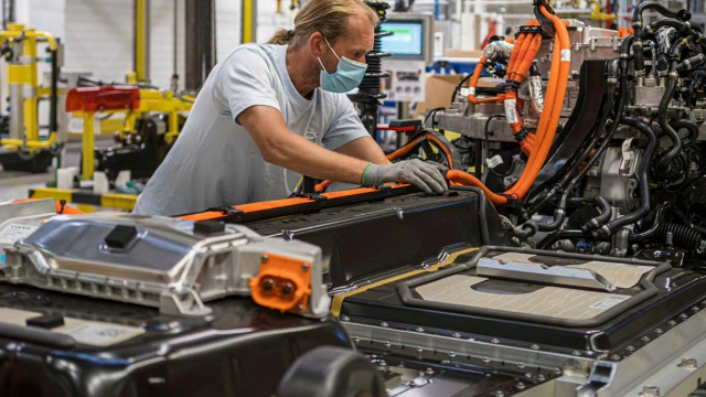 man-building-ev-battery-640x360 A man with a long blond ponytail leans over a long black box and wires on an assembly line in a warehouse.