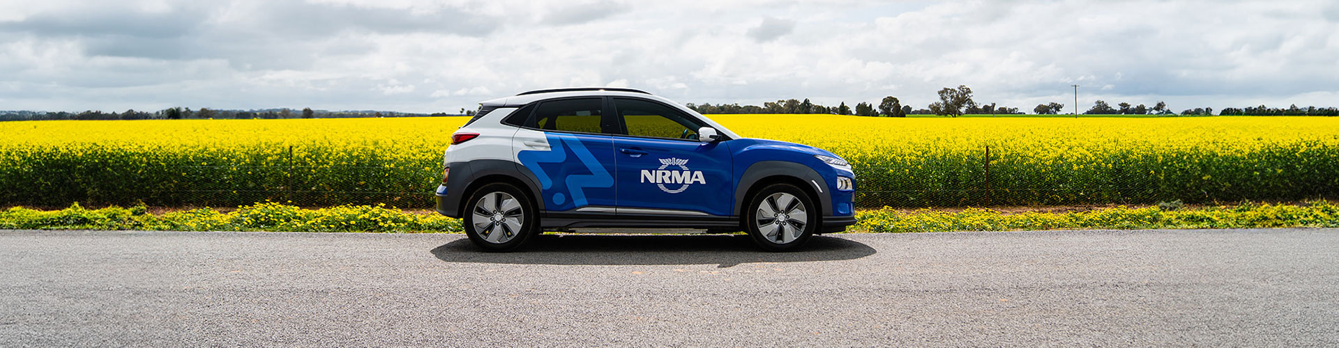 An electric car branded with NRMA logo parked in front of a field of canola