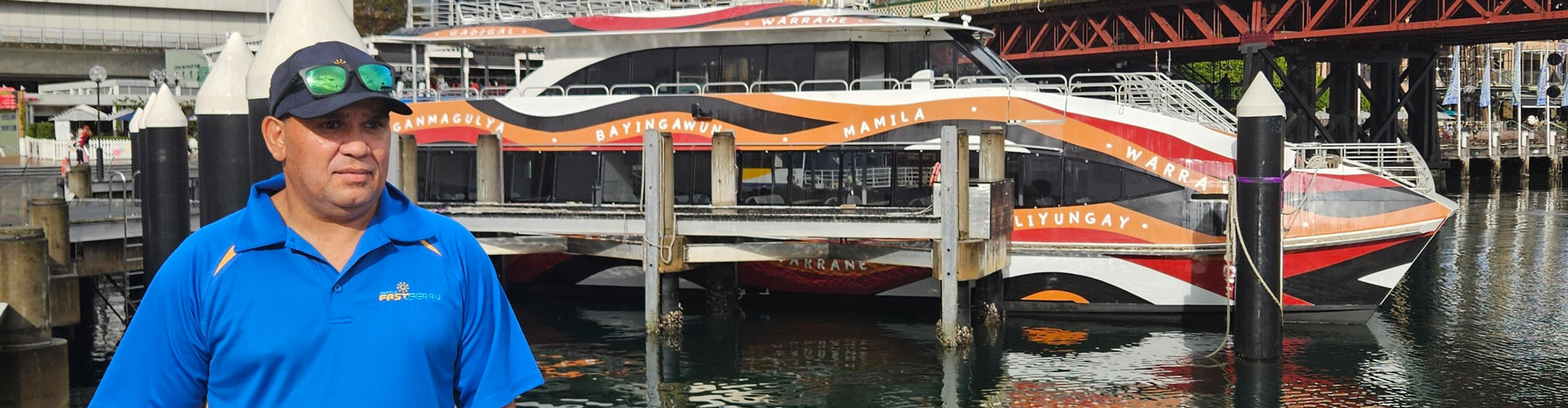 A man stands in a blue uniform with cap and glasses on his head, in front of a red and orange ferry