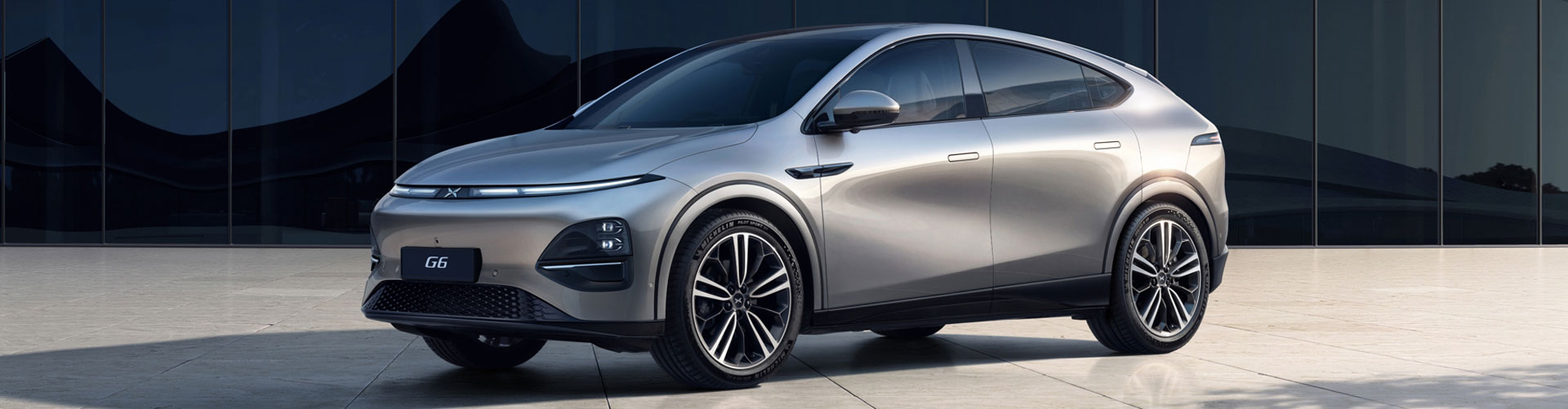 Silver Xpeng G6 parked om front of a large glass wall of a modern building