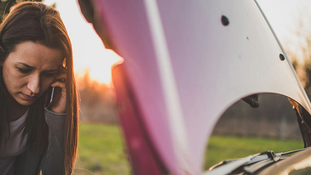 A woman on the phone looking into car engine at dusk