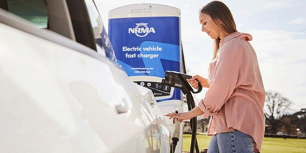A woman plugging a charger into the back of her car, under a sign that says, NRMA electric vehicle fast charger.