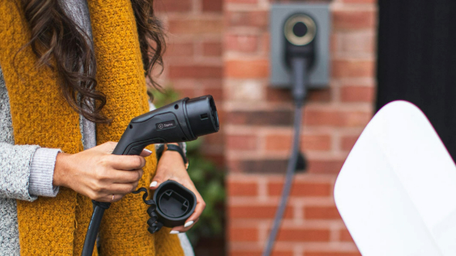 A woman's hand holding a shower nozzle shaped electric vehicle charger, about to plug it into a car.