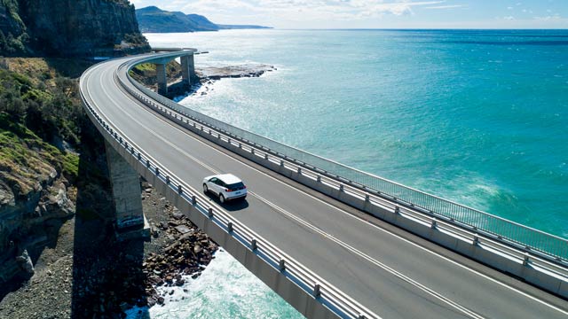 A car drives on Seacliff Bridge above aqua ocean 