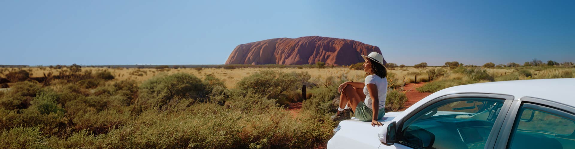 A woman sits on a car bonnet with Uluru in the distance