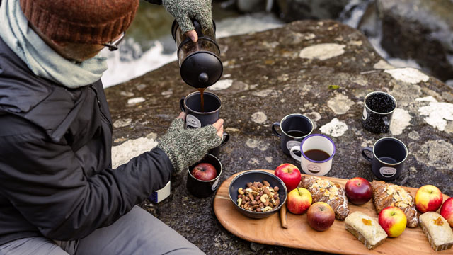 A man pours a hot drink sitting next to a rocky creek with a wooden board of apples and snacks