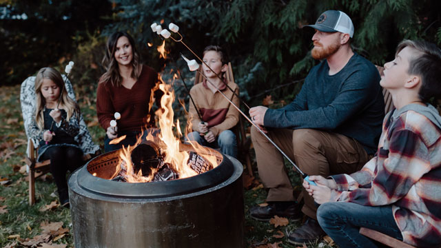 A family sits around a campfire in a copper brazier toasting marchmallows
