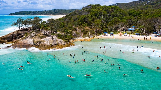 A rocky outcrop covered in trees creates a bay of turquoise water where people swim and kayak along the sandy beach tucked into the bay.