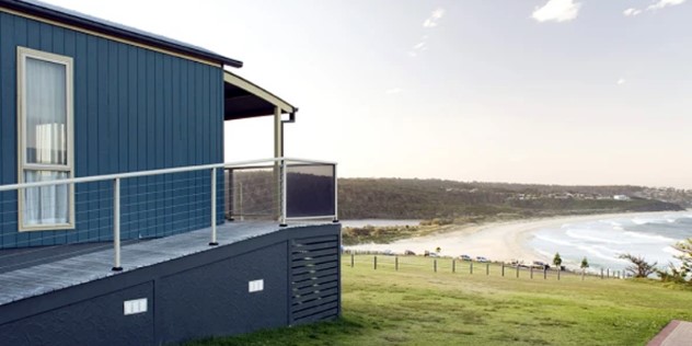 The veranda of a modern beach house overlooking a green hill leading down to a sunny beach and forested shore. 