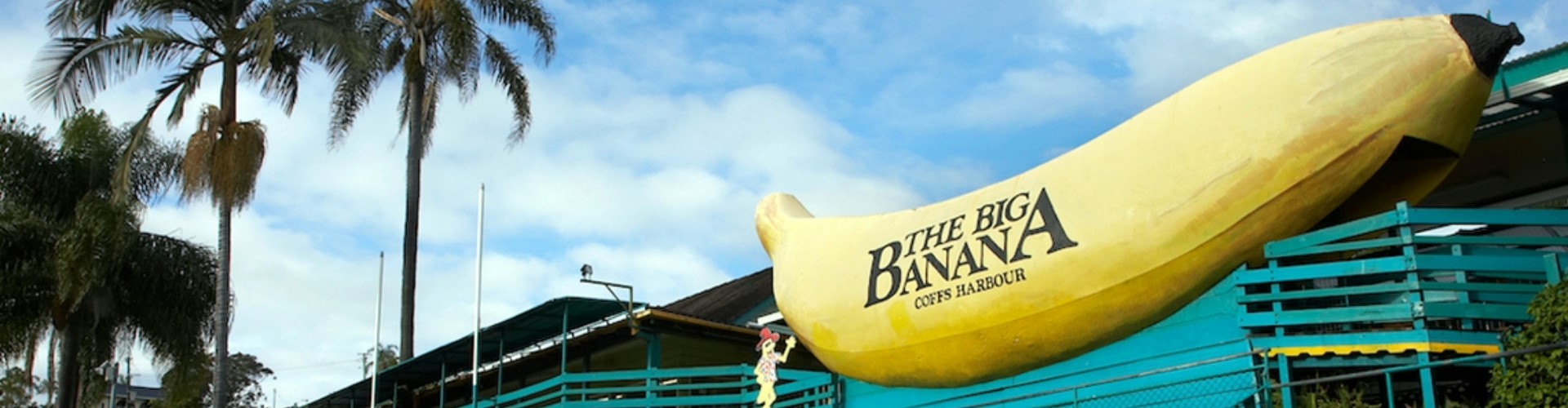 big-banana-nsw-1920x500 A giant yellow banana sculpture with the words The Big Banana, Coffs Harbour, written in black along the side, in front of a clubhouse and palm trees.