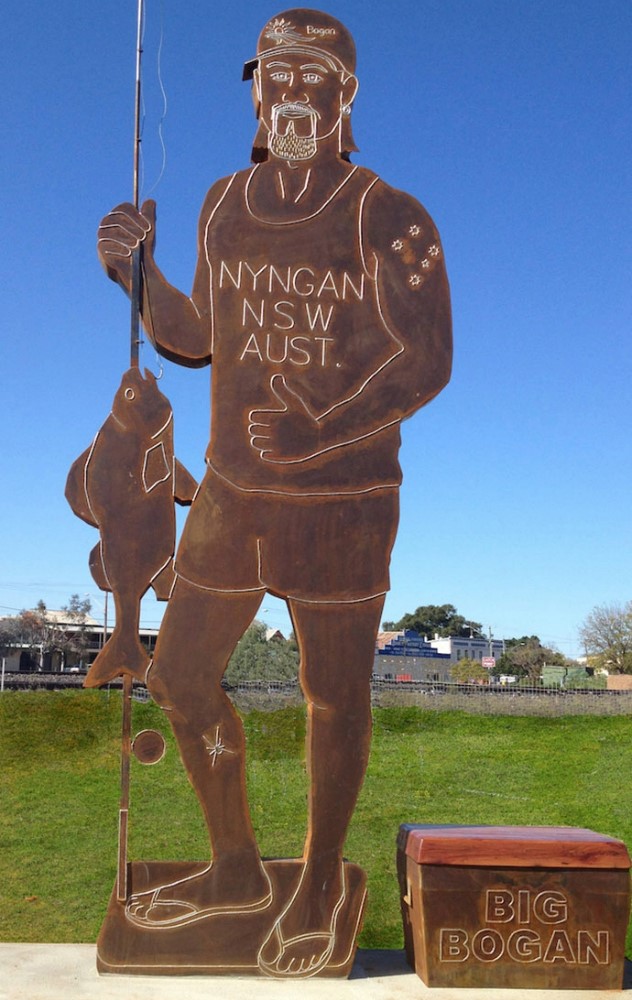 A flat, six metre tall brown metal sculpture of a man with a goatee and backwards cap wearing a tank-top, shorts and holding a fish while giving the thumbs up sign. Next to him is a square tackle-box with the words Big Bogan written on it. 