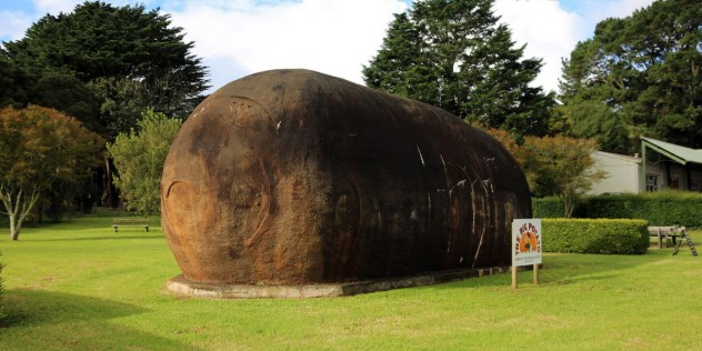 A ten metre tall sculpture of a brown potato laying on its side in a green garden on a sunny day. 